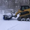 Yellow skid steer clearing snow with brush.